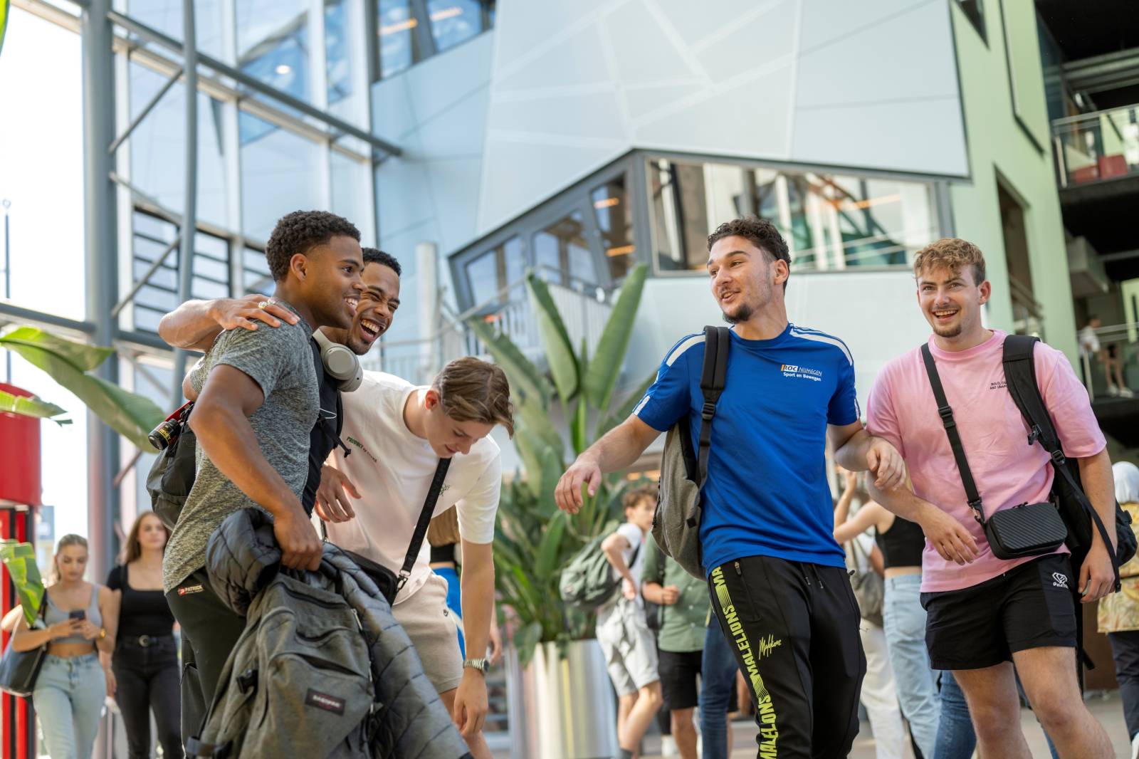 Vijf jonge mannen lachen en lopen samen door een lichte, moderne schoolhal.