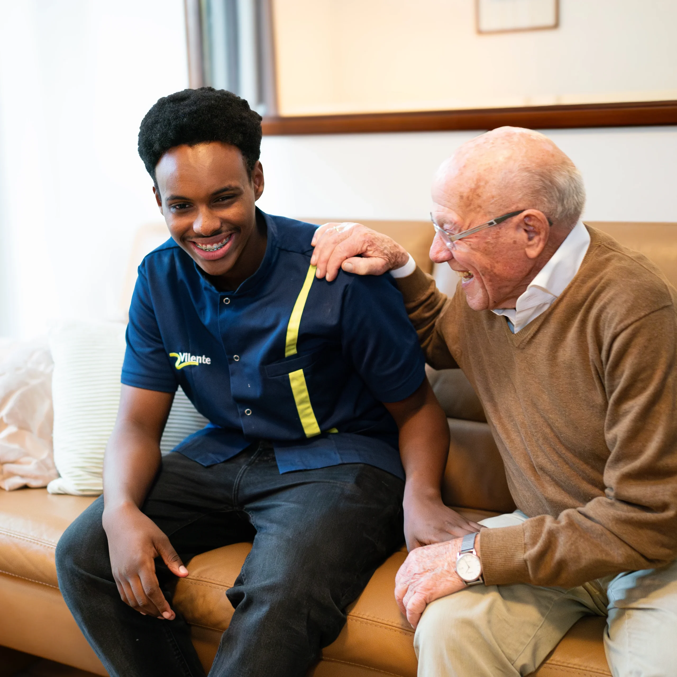 Oudere man en jonge zorgmedewerker lachen samen op de bank.