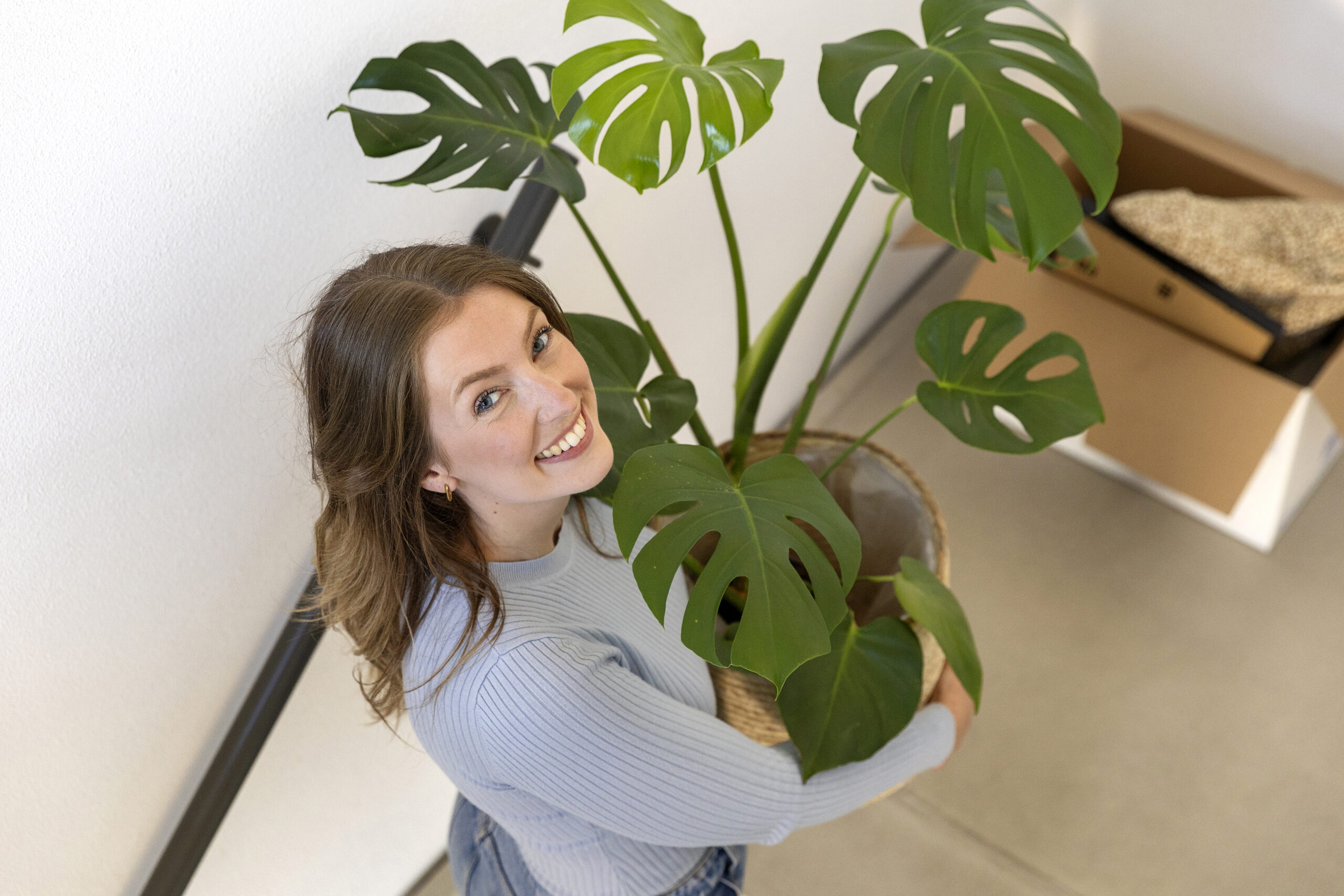 Vrouw lacht naar de camera met een grote plant in haar handen.